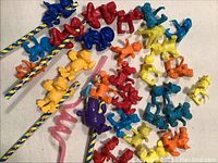 Colorful small plastic toy figures in red, blue, yellow, and orange on decorative sticks and straws. Multiple characters in various poses corresponding to popular franchises.