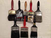 Photo showing seven paint brushes laid out flat, some packaged, some used, with a variety of bristle types and handle colors.