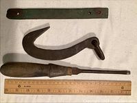 Four antique vintage tools arranged on a white cloth with a wooden ruler for scale, showing screwdriver, large meat hook, steel strip, and small hook