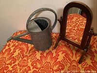 Photo showing a decorative metal watering can and a wooden swivel mirror on an orange patterned cloth
