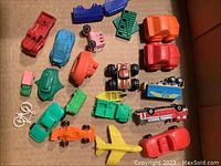 Top down view of a collection of small, colorful vintage plastic and rubber vehicles including cars, trucks, trains, a fire truck, a motorcycle, a bicycle, and an airplane arranged randomly in a box.