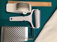 White plastic vegetable/pasta cutter with six blades, white lattice roller cutter, and metal four-sided grater, each placed alongside a ruler for size comparison.