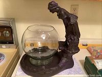 Side view of peasant lady figurine leaning over glass fishbowl mounted on a rounded base platform, showing details of figurine and fishbowl dimensions.