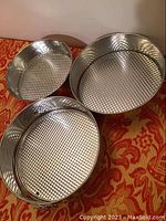 Photo showing three round metal springform pans with textured aluminum bases, nested inside each other on an orange-red patterned fabric.