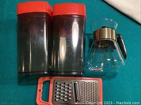 Two plastic storage bins with red lids, glass coffee percolator, and handheld grater laid out on a surface.