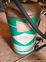 Full view of vintage sprayer showing rusted metal lid, green body with white label and attached hose.