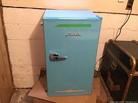 Frontal view of robin's egg blue RCA bar fridge against a basement wall, showing chrome handle and RCA logo.