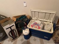 Photo showing blue and white Igloo cooler open containing paper plates and a white container, with blue Playmate cooler jug in front and cardboard boxes behind.