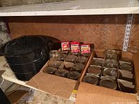 Wide view of the black enamel canning pot, boxes of clear glass canning jars with metal lids, and boxes of Ball brand metal rings on a basement shelf.