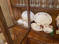 Glass cabinet display of 12 Royal Albert bone china tea cups and 2 platters with pink rose pattern and gold trim.