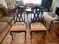 Two vintage wooden dining chairs with upholstered beige seats and carved backrests, placed side by side in a living room.