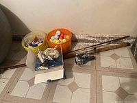 Full view of fishing gear lot showing two fishing rods, two reels (one boxed), yellow bucket with fishing items, orange bucket with golf balls on tiled floor.