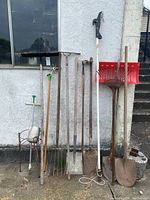 Photo of various garden tools including multiple shovels, spades, rakes, a branch lopper, and a plant stand outside against a wall.
