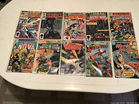 Top-down view of 10 assorted comic books laid out on a white table, each in plastic protective sleeves. Covers visible with multiple Marvel and DC titles.