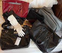 Multiple purses and gloves arranged on a white surface showing different styles and materials. Purses include black leather, faux reptile skin, patent red, and metallic gold clutch. Gray carry-on bag is visible at the back.