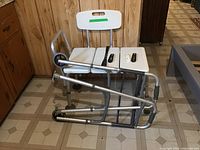 White plastic shower bench with three seats, metal frame with rubber feet, placed next to a folded walker indoors.