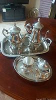 Full view of silver plated coffee pot, tea pot on large rectangular tray, cream pitcher and sugar bowl on smaller oval tray, displayed on wooden table with some reflections and tarnishing visible