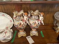 Pair of ornate Capodimonte porcelain vases with cherub figures displayed on a wooden shelf with other ceramics.