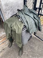 Green Pro Tac waders with attached boots and green rain jacket placed on a crate outdoors.