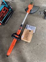 Black & Decker orange and black electric weed eater laid out on concrete floor near red and blue lawn tool.