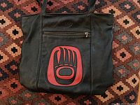Front view of the black leather handbag with red Indigenous-style bear paw design on front and external zipper pocket.