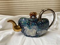 Side view of teapot showing glazed body with foliage and white cat motif, gold-tone spout, silver-tone handle and apple-shaped lid finial