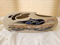 Full front view of elongated ceramic vase showing carved top cutouts and glazed wave and landscape design on the side.