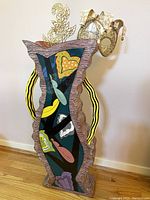 Full view of large uniquely shaped pottery ceramic vase with colorful abstract painting and decorative mask on top, against wall and hardwood floor.