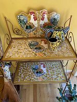 Full view of metal baker's rack showing three mosaic-tile shelves with rooster motifs, plus ceramic teapot and mug on top shelf.