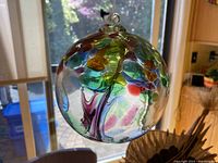 Glass art orb suspended in front of a window, showing colorful internal tree design with leaves in green, blue, yellow, and orange colors. The clear glass orb hangs by a small top loop.