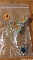 Photo showing two brooches: a butterfly enamel and rhinestone piece and a floral rhinestone brooch with blue center stone, both placed on wood surface inside a plastic bag with orange sticker.