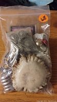Photo of a sealed plastic bag containing various assorted jewelry items including a silver-tone metal cuff bracelet, beads, earrings, and a circular cream-colored decorative piece.