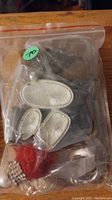 The lot of assorted jewelry in a clear plastic bag. Visible are a large oval silver-tone pendant or brooch, two smaller matching silver pieces, red and white faux pearl strands, and rhinestone jewelry.