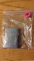 Silver-tone brushed metal Zippo lighter in a plastic bag on a wooden surface.
