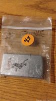 Photo of a silver metal Zippo lighter with an engraved image of a polo player on horseback, placed inside a resealable plastic bag on a wooden surface.