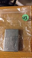 Photo shows a used metal Zippo lighter in a plastic bag with a green sticker labeled '42' on a wooden surface.