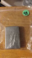 Photo of a silver-tone metal Zippo lighter with a brushed finish on a wooden surface inside a plastic bag.