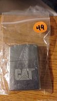 Photo of a silver-tone metal Zippo lighter featuring the CAT logo, inside a clear resealable plastic bag with an orange sticker marked 49.