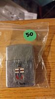 Photo of a silver-tone Zippo lighter with a colored shield crest emblem on front showing wear and scratches, enclosed in a plastic bag with a small green sticker labeled '50'.