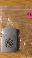 Photo shows a silver-tone metal lighter with a raised eagle emblem on the front, placed on a wooden surface inside a clear plastic bag.
