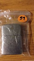 Metal Zippo-style lighter in a plastic bag with a sticker '57'. Scuffs and scratches visible on surface.