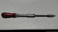 Manual hand drill placed on a flat surface showing wooden handle, textured metal shaft, and bit holder with a screwdriver bit attached.