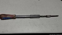 Handheld screwdriver with wooden handle, telescoping metal shaft, interchangeable flathead bit attached, placed on white surface.