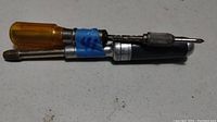 Photo showing a manual screwdriver with an amber plastic handle and two bits attached: a drill bit and a Phillips screwdriver bit, secured with blue tape.