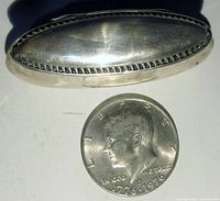 Top view of the oval sterling silver pill casket beside a 1776-1976 Kennedy half dollar coin for size comparison, showing the polished smooth surface and raised edge detail.