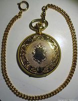 Closed gold tone pocket watch showing front case with ornate shield design and attached gold tone chain.