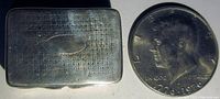 Top view of rectangular silver pill box with engraved text and oval cartouche next to a coin for scale.