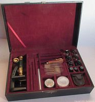 Open wooden case showing vintage brass microscope on the left, various glass slides in plastic holders, small bottles, test tube, round petri dishes, pipette and spring magnifier organized inside red velvet lining.