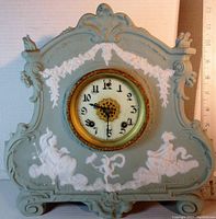 Front view of green jasperware mantle clock showing white cameo figures and floral garlands with clock face at center.