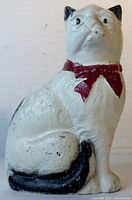 Side angle showing the white cast iron cat with black ears, tail, and paws, and red ribbon collar.